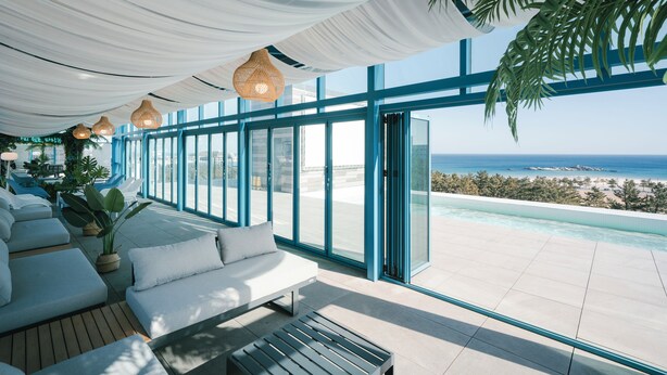 Un área lounge moderna al aire libre con vista al océano, que incluye un sofá, una mesa de madera e iluminación colgante.