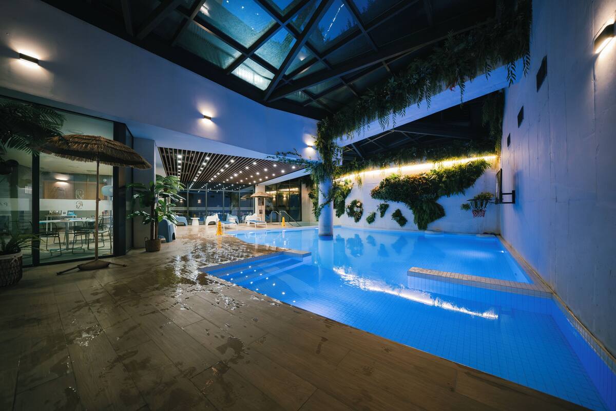 An indoor swimming pool with a glass ceiling, surrounded by indoor seating and greenery.