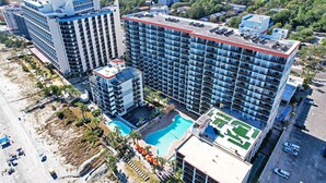Un hotel moderno de gran altura con varias plantas, una piscina en la azotea y una zona de playa con sombrillas.