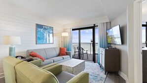 Habitación de hotel con sofá, sillas, una mesa de centro, televisor y balcón con vistas.