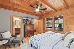 A cozy bedroom with a wooden ceiling, a bed with a patterned blanket, a dresser, and a window with a view of the outdoors.
