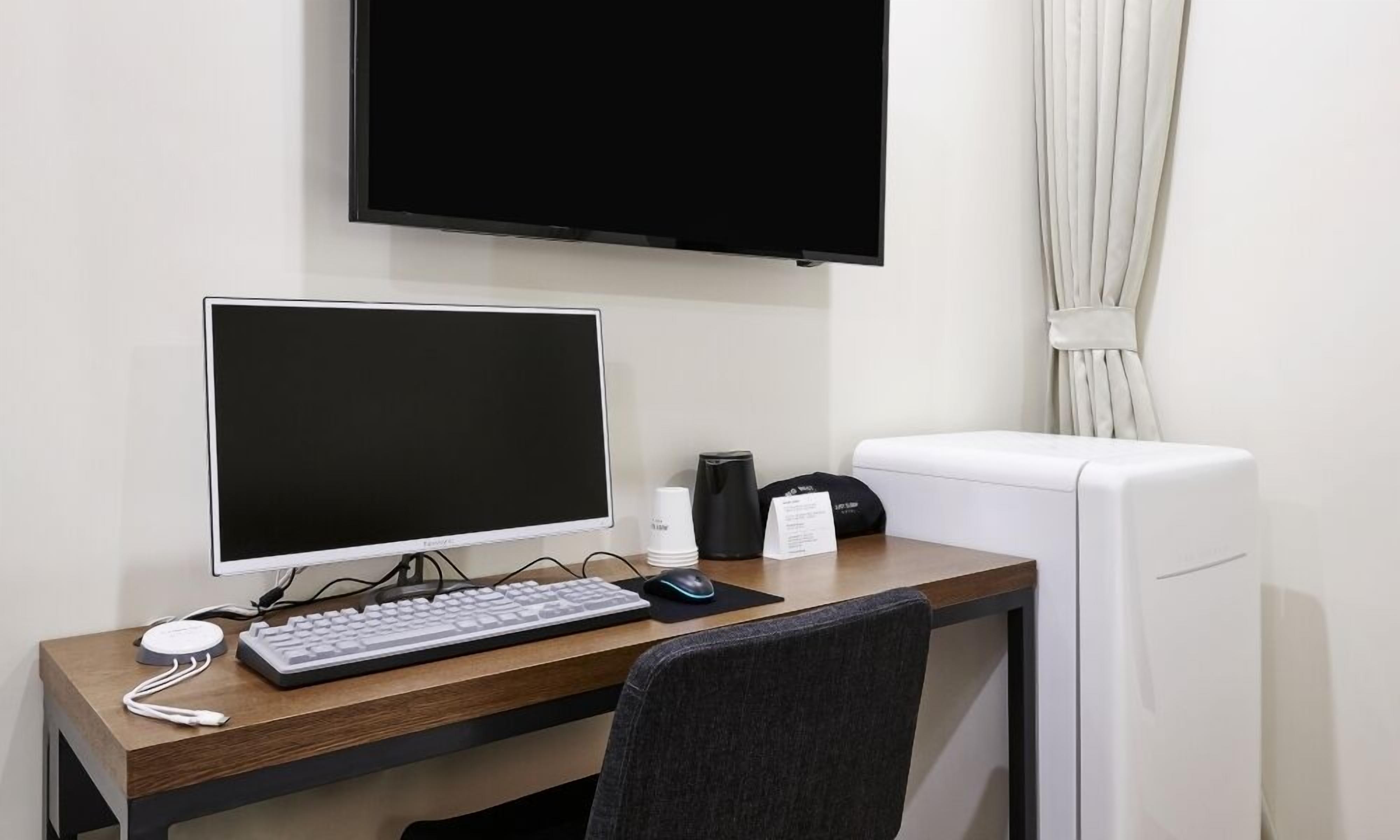 Une chambre d’hôtel équipée d’un bureau en bois, d’un ordinateur de bureau, d’un clavier, d’une souris, d’un écran, d’une machine à café et d’un réfrigérateur blanc.