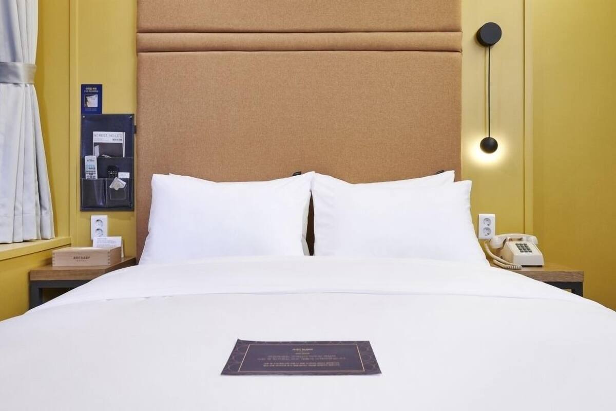 Habitació d'hotel amb llit, almoines blanques, una taula de nit amb telèfon, una làmpada penjada a la paret i una finestra amb cortines.