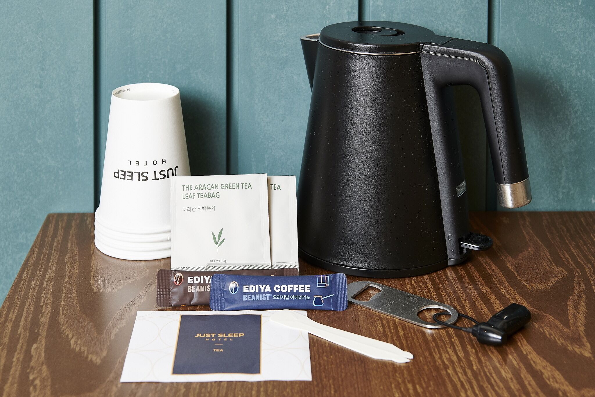 A coffee maker, a cup, a tea bag, a packet of coffee, and a spoon on a wooden table.