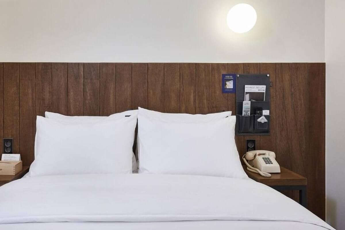 Habitació d'hotel amb capçal de llit de fusta, llit amb roba de llit blanca, una tauleta de nit amb una caixa de mocadors, un telèfon i un panell de control fixat a la paret.