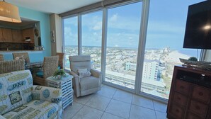 A spacious living room with a large window offering a city view, wicker furniture, and a television on a wooden cabinet. - Island Tower 1501 - Right on the Beach - Corner (Gulf Shores)