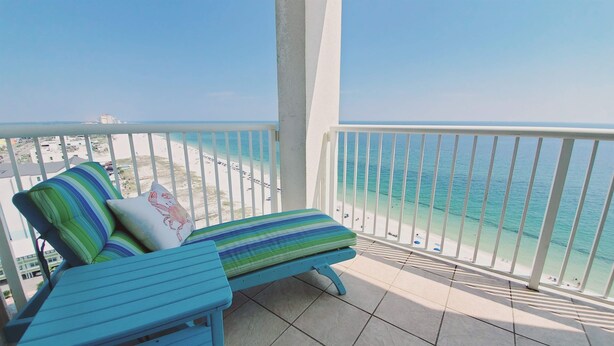 A balcony with a blue lounge chair and cushions, overlooking a beach with clear turquoise water and a sandy shore. - Island Tower 1501 - Right on the Beach - Corner (Gulf Shores)