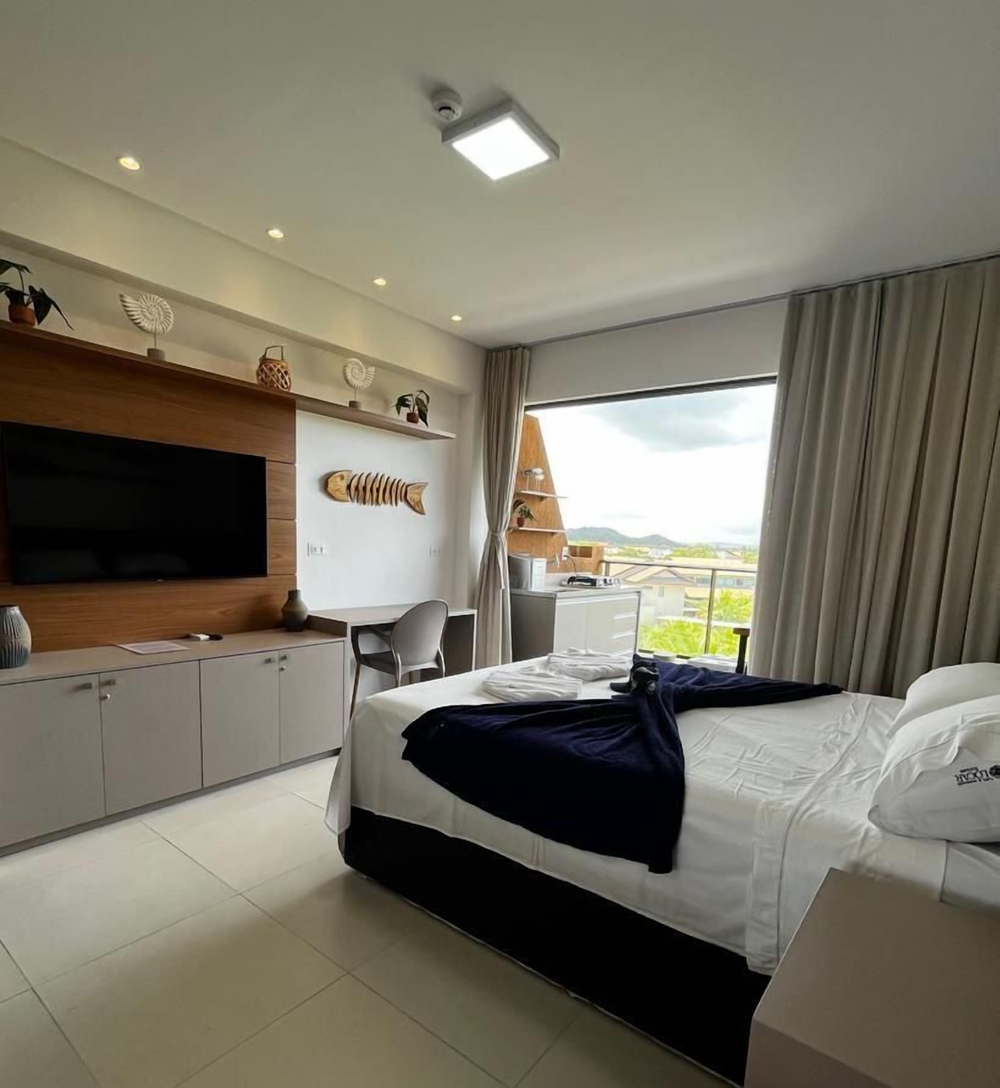 A modern hotel room with a large bed, a flat-screen TV, a desk with a chair, and a view of the outdoors.