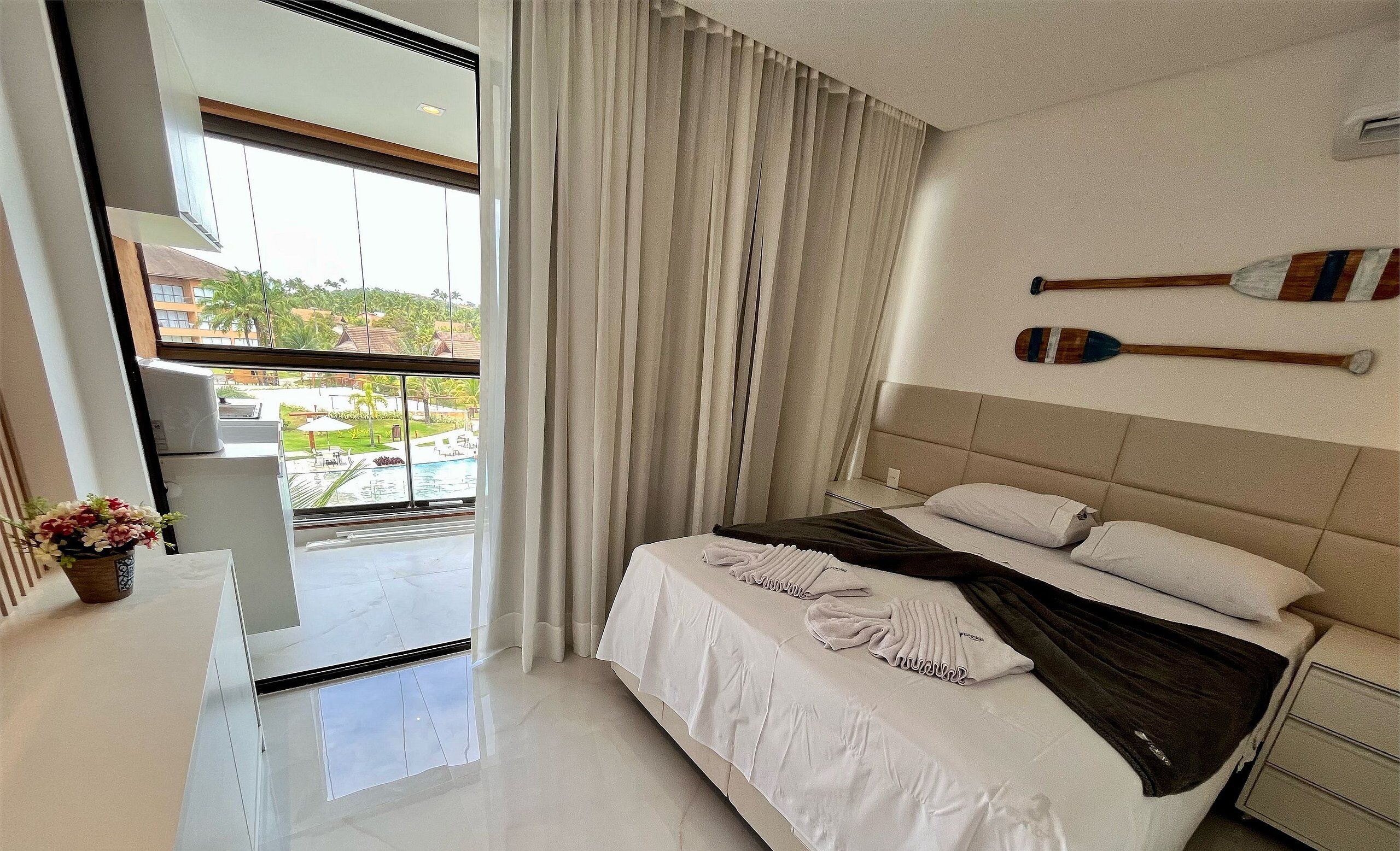 A hotel room with a bed, a nightstand, a window with curtains, a view of a pool and buildings, and a wooden paddle on the wall.
