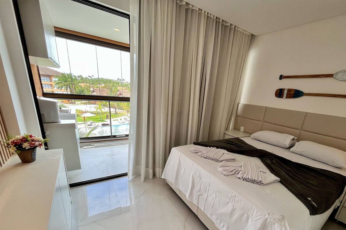 Ein Hotelzimmer mit einem Bett, einem Nachttisch, einem Spiegel, einem Fenster mit Vorhängen und Blick auf einen Pool sowie Gebäude.