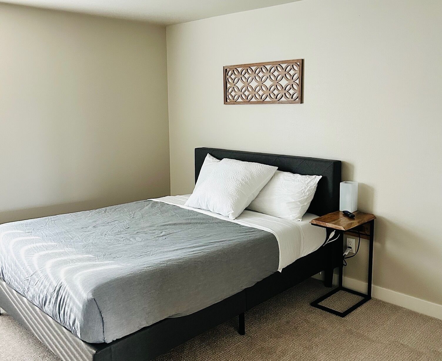 Cama bem arrumada com colcha cinza e almofadas brancas, mesa de cabeceira com abajur, e um objeto decorativo na parede acima da cama.
