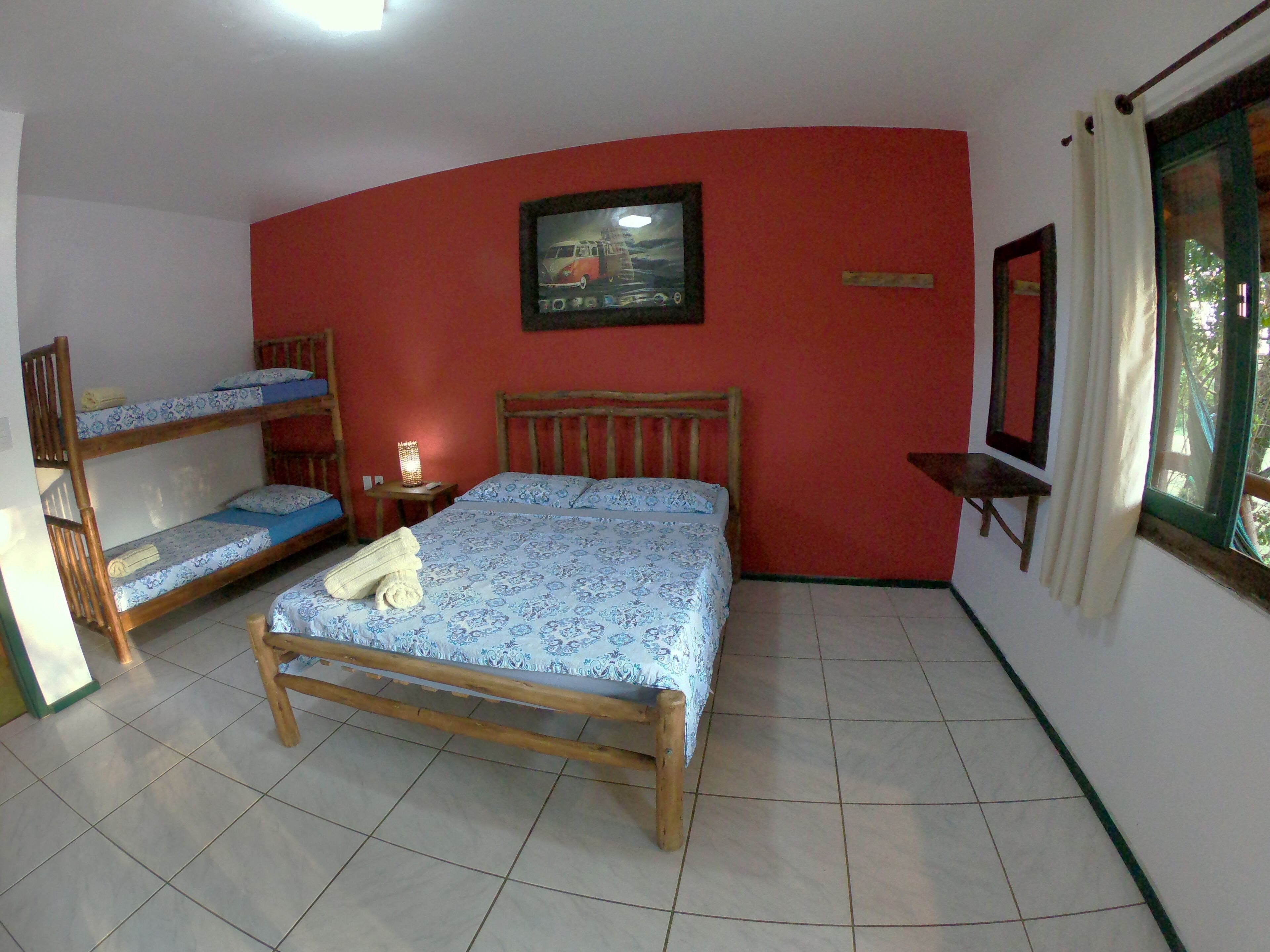 A room with a bunk bed, a single bed, a bedside table with a lamp, a framed picture on the wall, and a window with curtains.