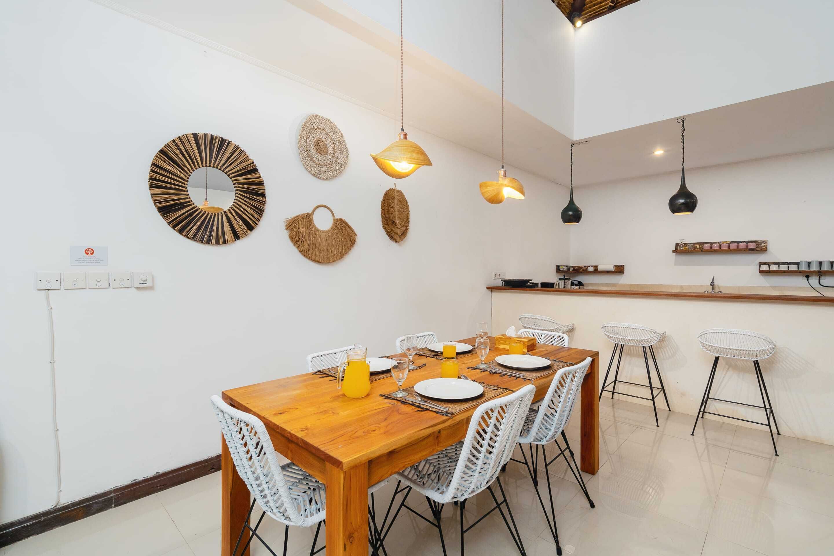 Un coin repas avec une table en bois dressée pour quatre personnes, des chaises blanches et des luminaires suspendus.