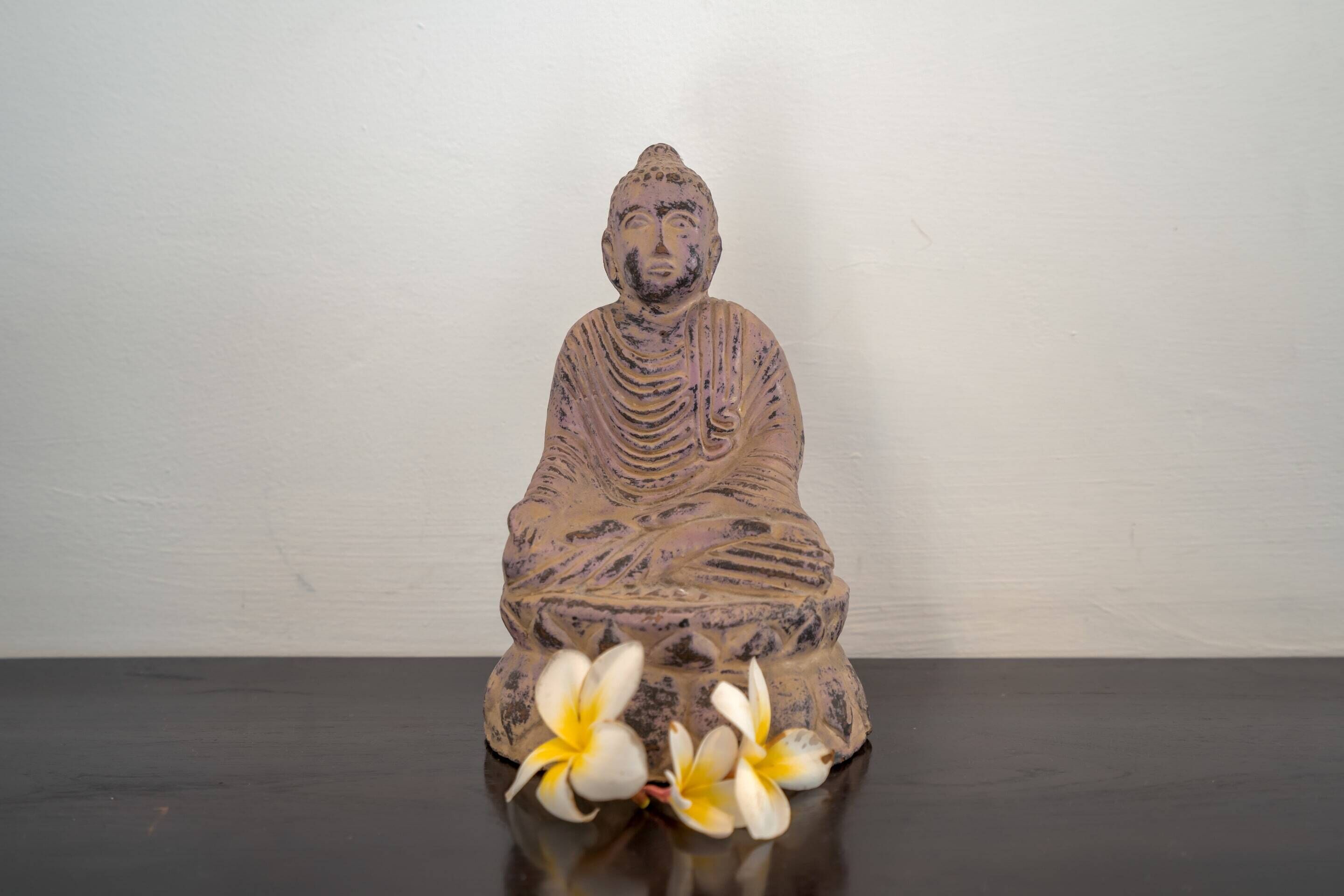 Une statue représentant une personne assise, vêtue d’une robe drapée, placée sur une surface sombre, avec deux fleurs devant.