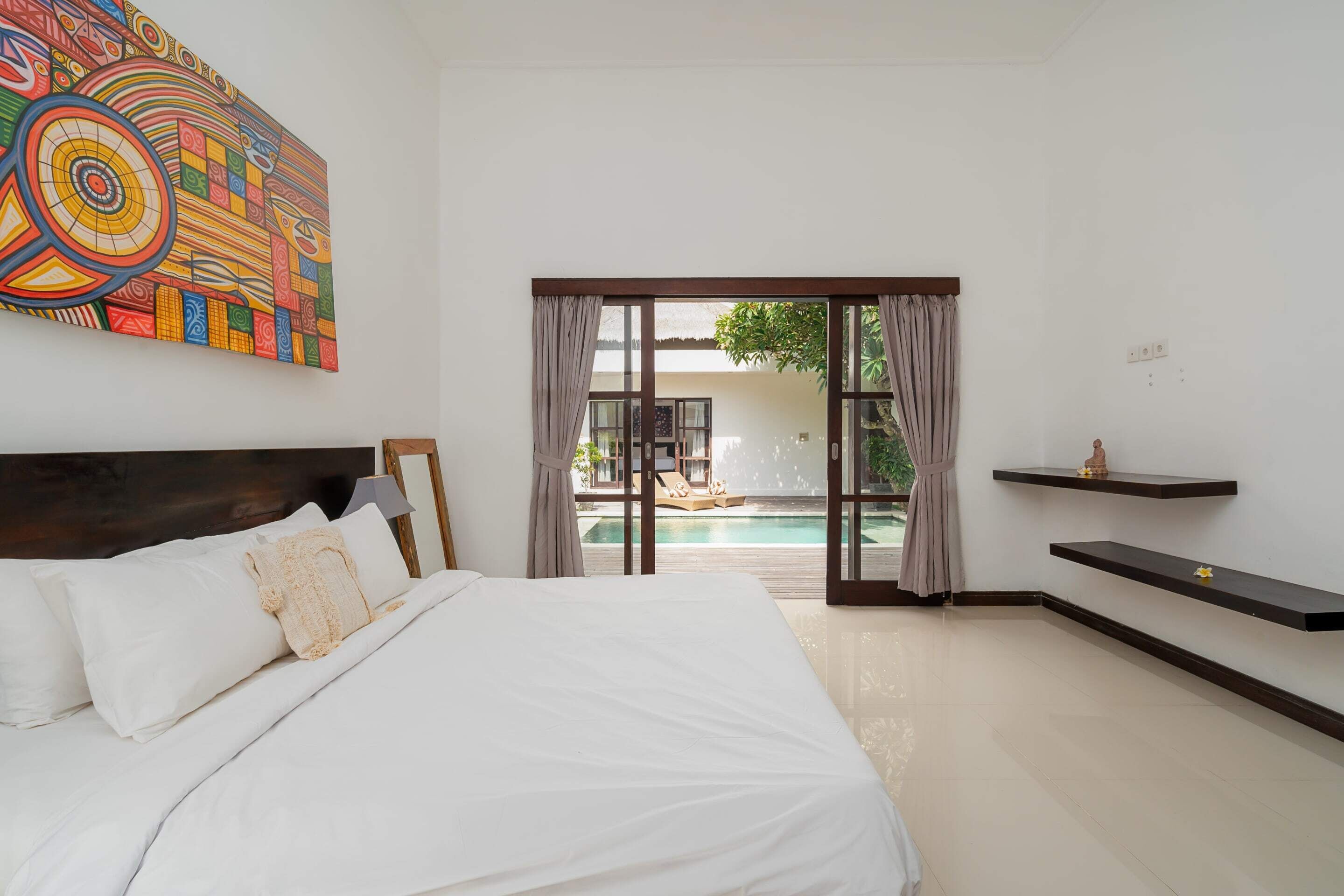A bedroom with a large bed, a colorful abstract painting, a view of a pool, and a sliding glass door.