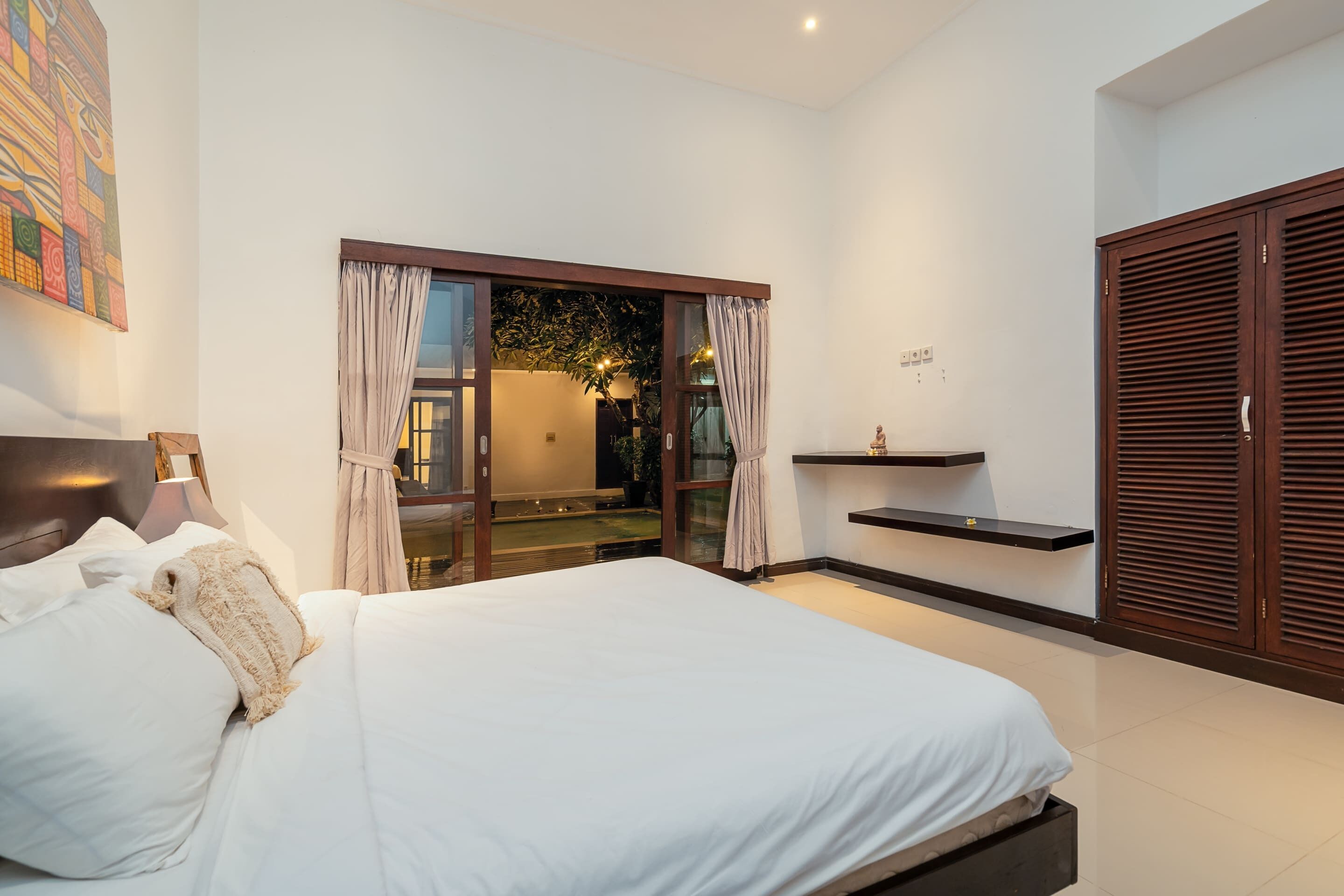 Une chambre moderne avec un grand lit, une porte-fenêtre coulissante donnant sur un balcon, une petite étagère et une armoire.