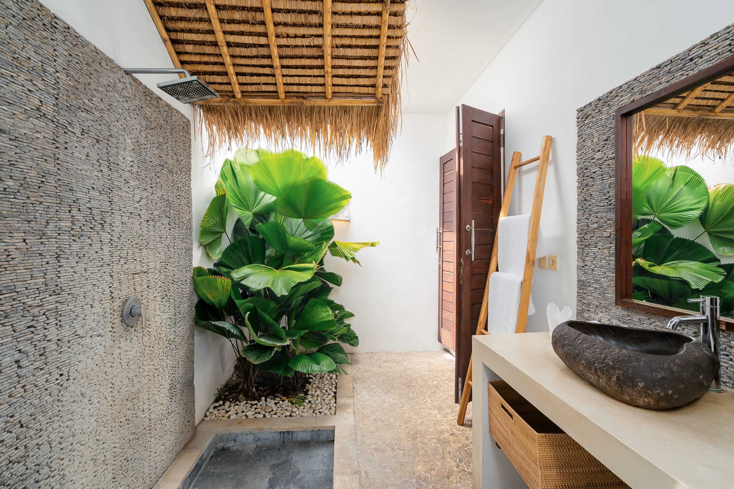 A bathroom with a stone wall, a large mirror, a stone sink, and a shower with a thatched roof.