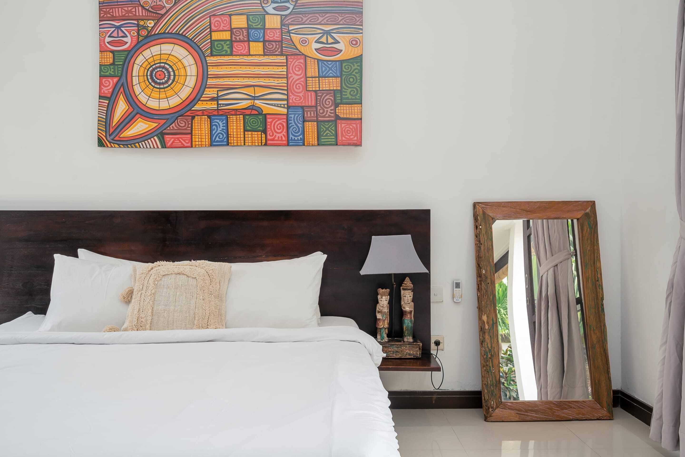 A neatly made bed with white linens, a wooden headboard, a colorful abstract painting above the bed, a nightstand with a lamp and a statue, and a large mirror leaning against the wall.