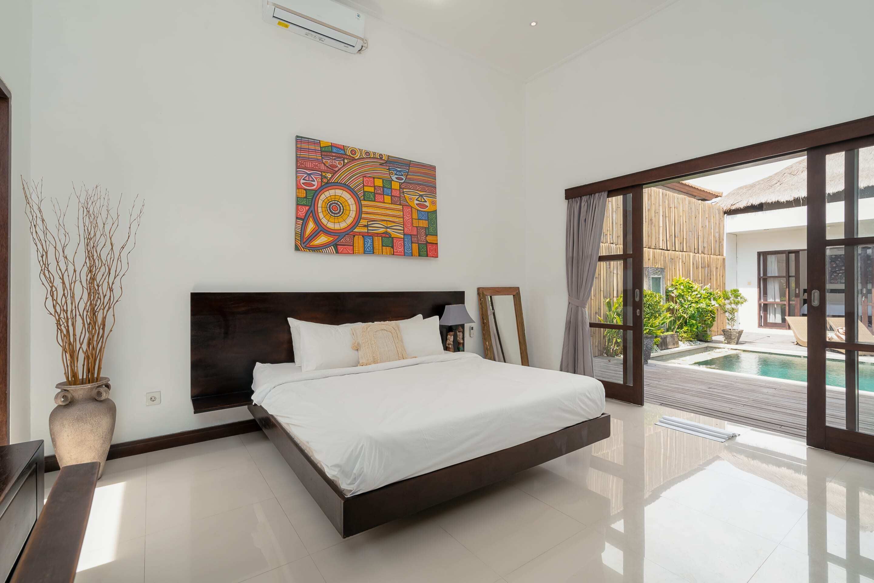 A modern bedroom with a large bed, a decorative wall piece, and a view of an outdoor swimming pool.