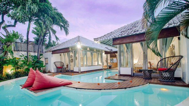 Piscina con cubierta de madera y cojines rojos, rodeada de plantas tropicales y un área de estar techada con sillas de mimbre.