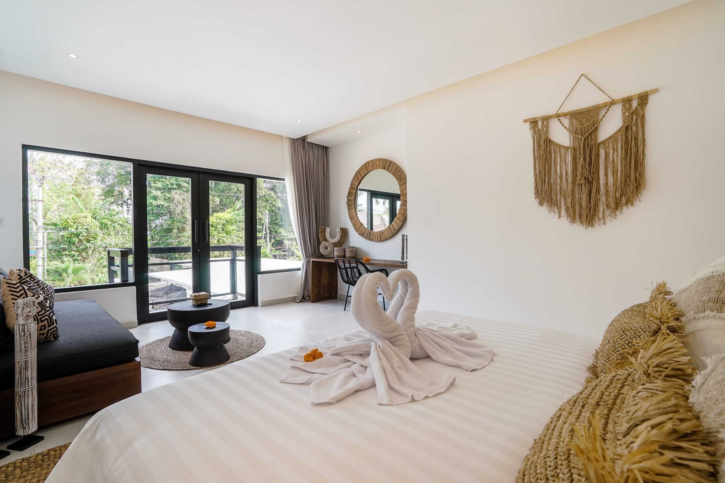 A modern bedroom with a large bed, a sofa, a round mirror, and a view of greenery outside.