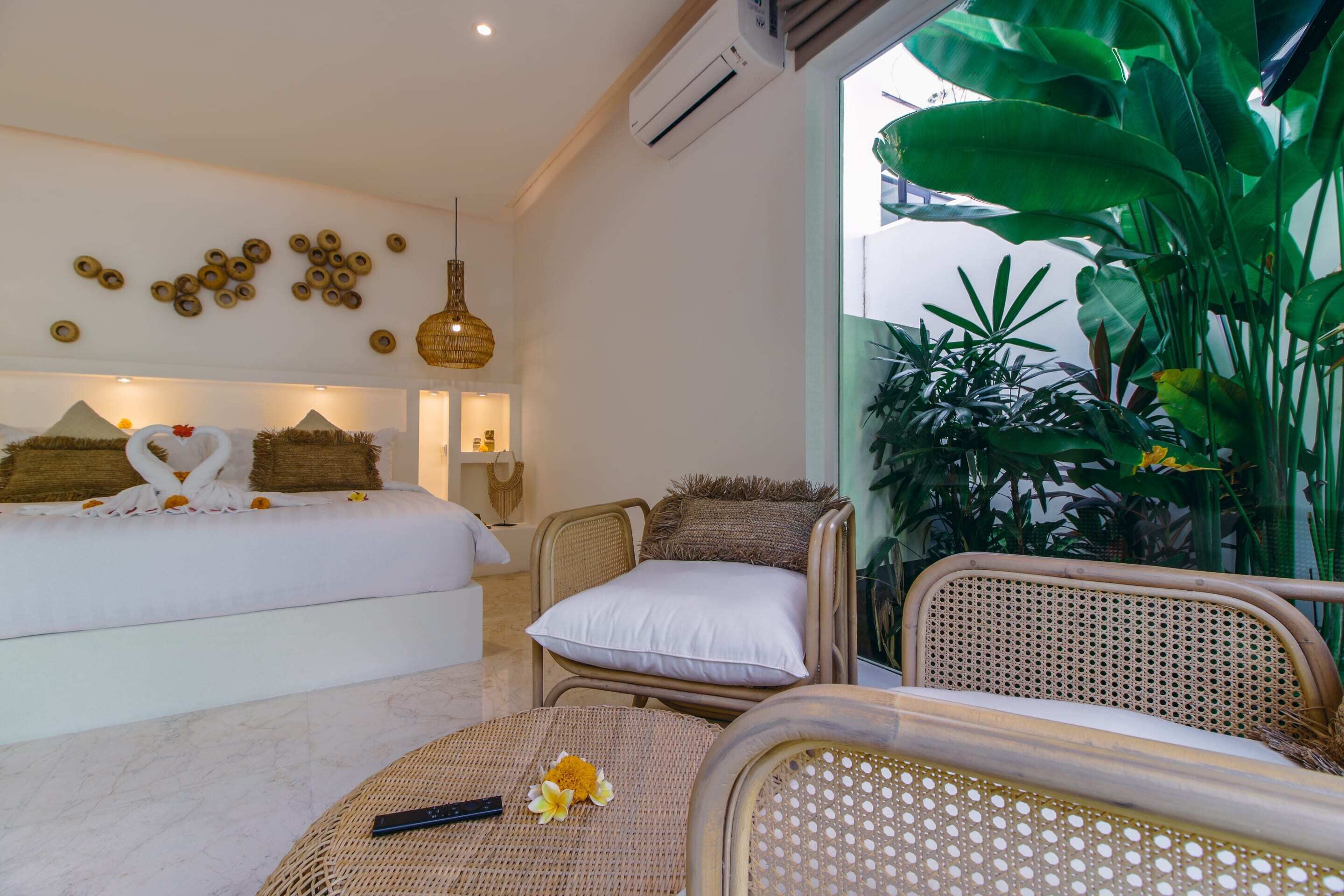 A hotel room with a bed, a wicker chair, a round table, and a large plant.