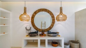 Uma toucador de banheiro com um espelho redondo, duas luminárias pendentes e uma prateleira com objetos decorativos.
