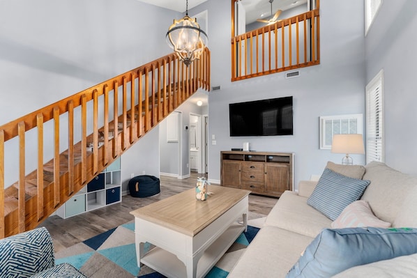 A modern living room with a wooden staircase, a flat-screen TV mounted on the wall, a wooden cabinet, and a beige sofa with blue and white cushions.