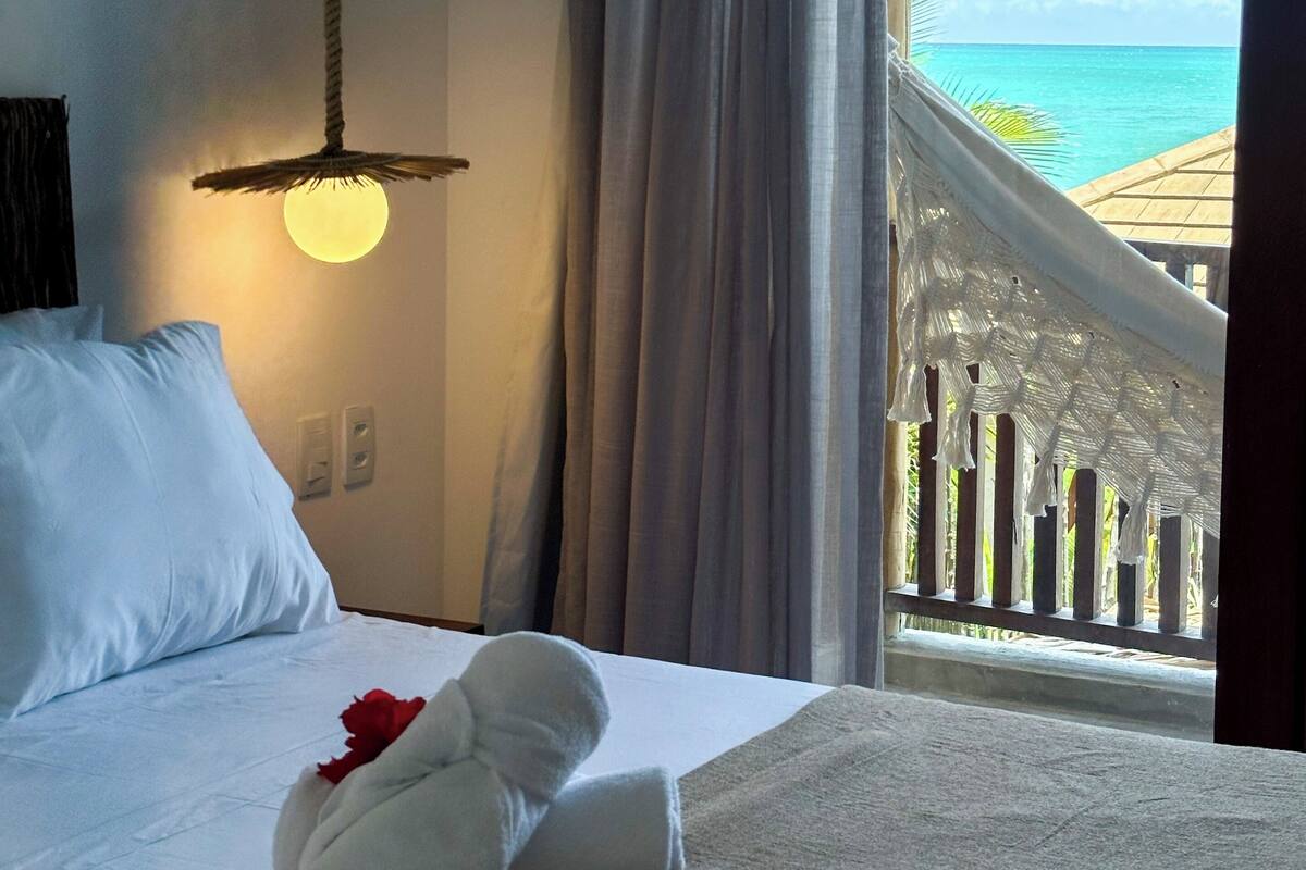 Quarto de hotel com cama, luminária pendente, vista para o oceano e varanda com guarda-corpo.