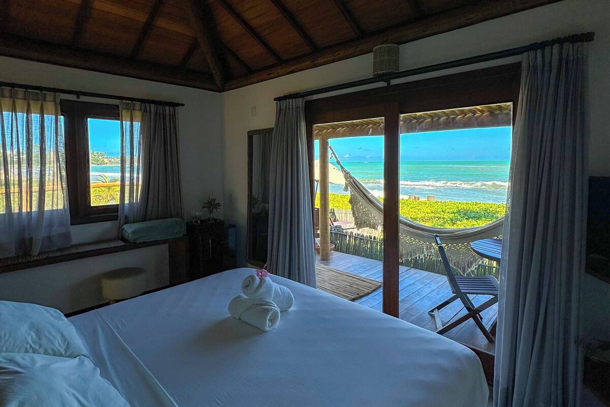 Quarto com cama, vista para o mar e varanda com mesa e cadeiras.