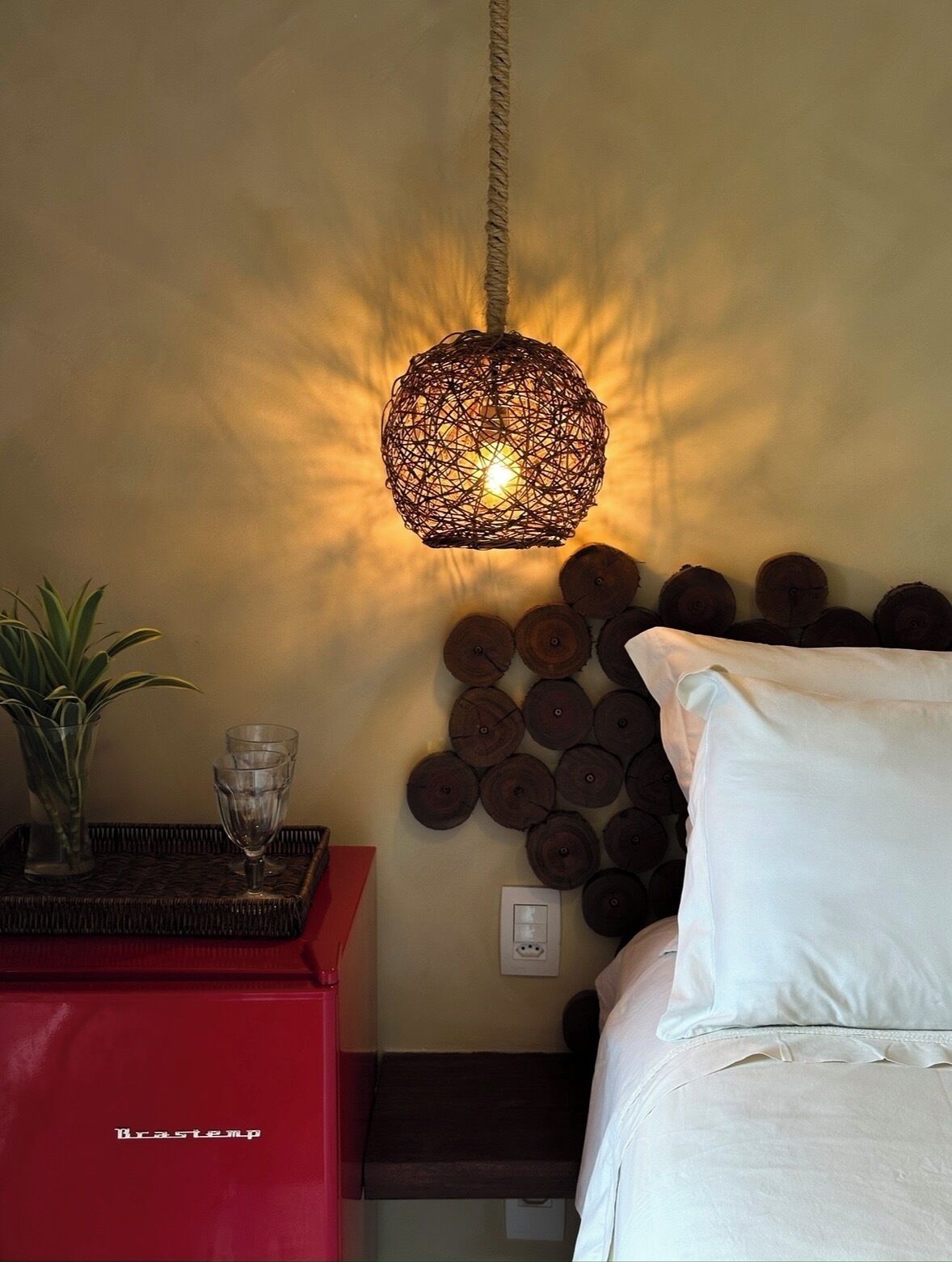 Une chambre avec un luminaire en fibres tressées, une décoration murale en forme de bûche en bois, une table de chevet avec un verre et une armoire rouge.