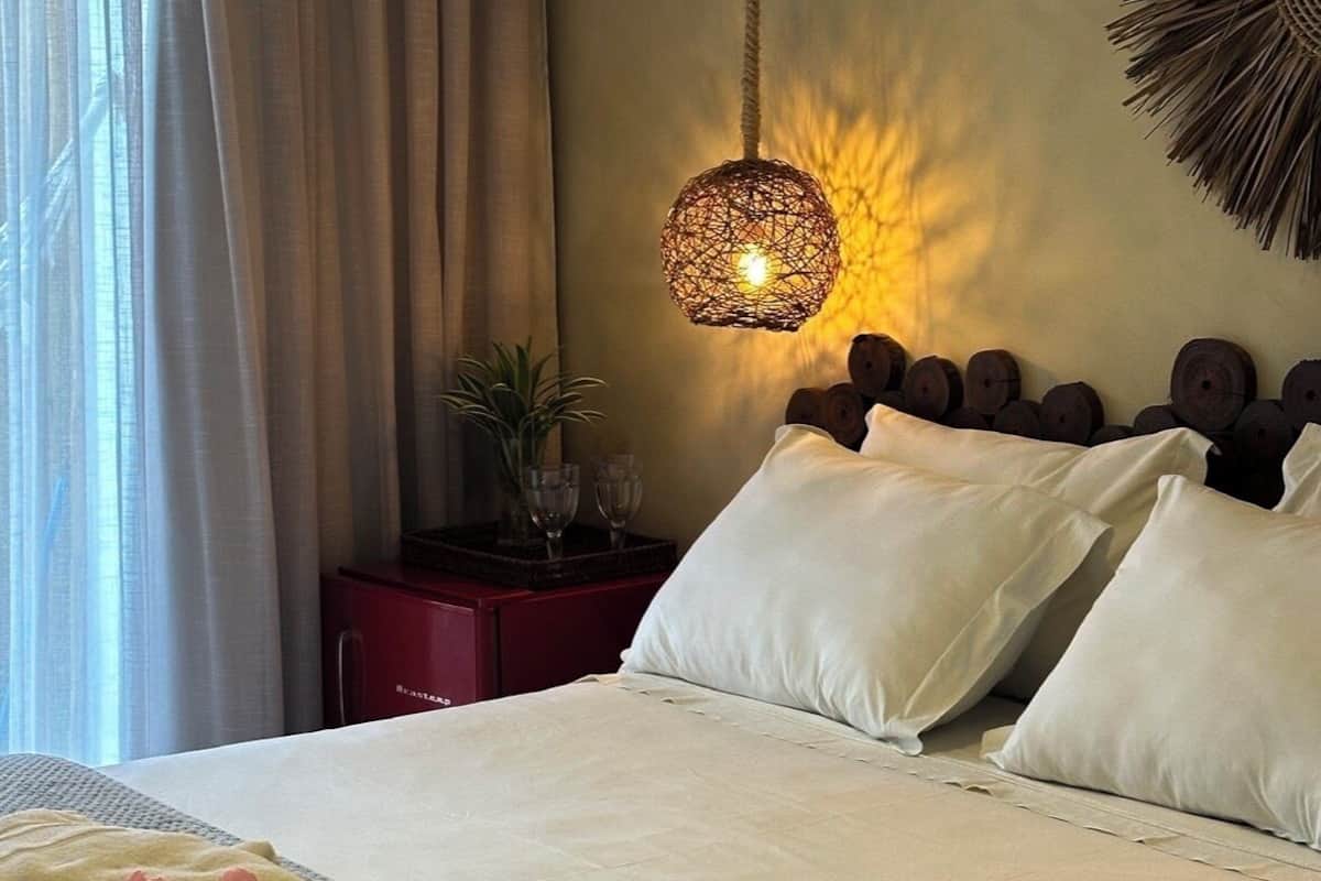 Quarto com cama, almofadas, mesa de cabeceira com copos, luminária pendente e um enfeite de parede.