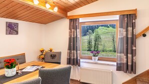 Un comedor con una mesa de madera, sillas grises, un florero con flores rojas y una ventana con cortinas.