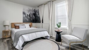 A bedroom with a bed, a chair, a window with curtains, a wall-mounted painting, and a small table with a plant. - Modern Zen Farmhouse to help you recharge and rejuvenated (Royal Oak)