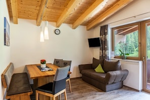 Ein gemütliches Zimmer mit Holzbalkendecke, einem Esstisch mit Stühlen, einer Couch mit Kissen, einem Fernseher und einem Fenster mit Blick auf Bäume.