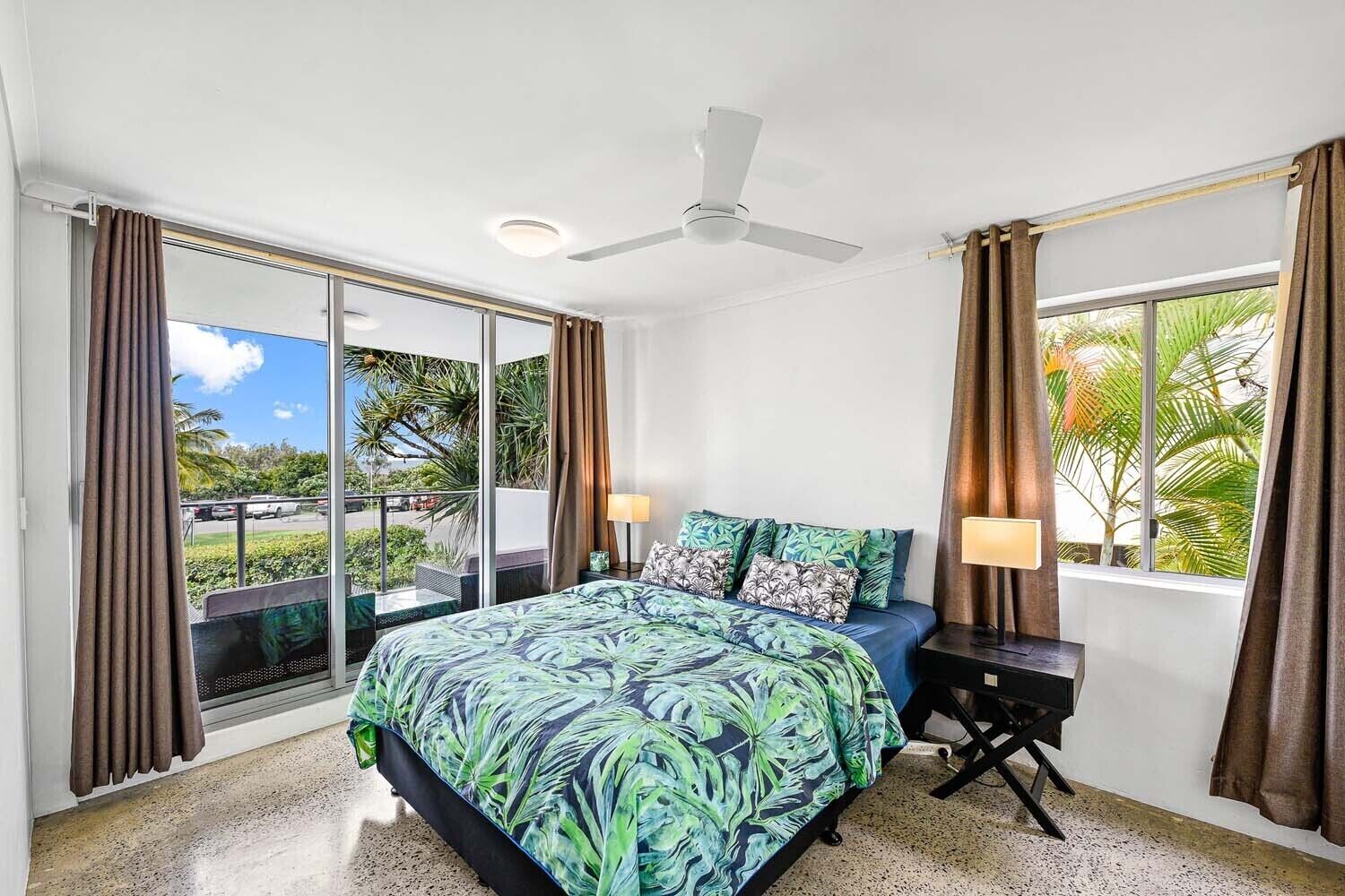 Habitación de hotel con cama, ventilador de techo y vista a palmeras y un estacionamiento.