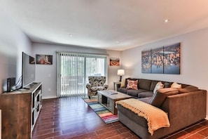 A modern living room with a sofa, armchair, and coffee table. There is a TV on a wooden stand, a window with blinds, and artwork on the wall.