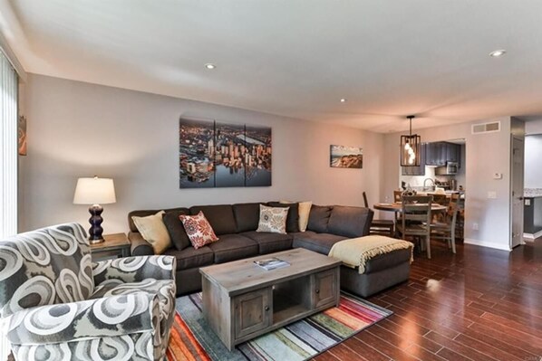 A modern living room with a sectional sofa, a patterned armchair, and a coffee table. There is a cityscape painting on the wall, a dining area with a chandelier, and a kitchen in the background. - 2 Bed, 2.5 Bath Lake Murray Townhouse Loaded With Amenities! (La Mesa)