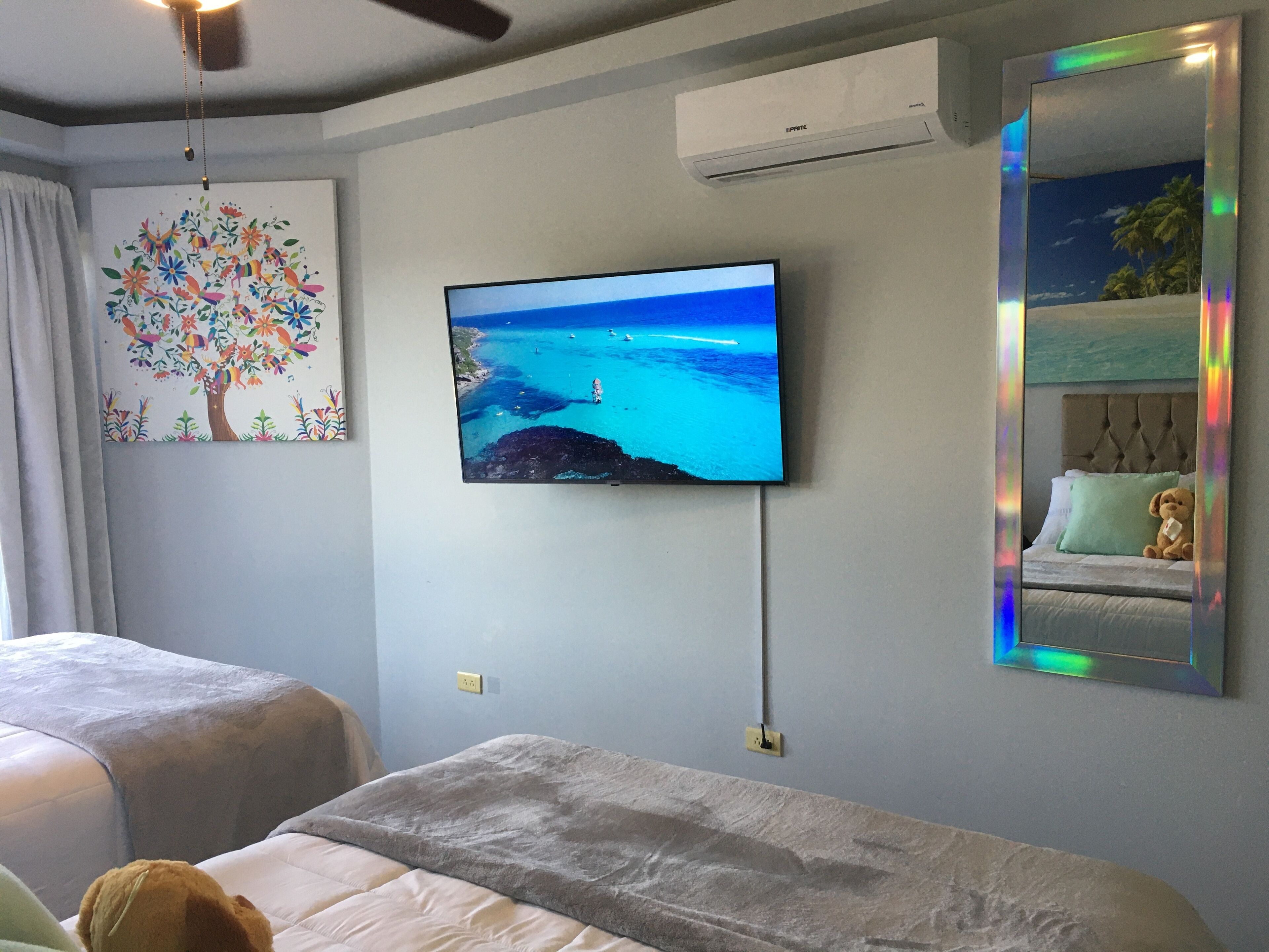 A bedroom with a TV mounted on the wall, a bed with a grey duvet, a decorative tree painting, and a ceiling fan.