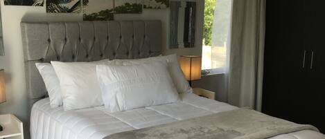 A bedroom with a large bed, a headboard, a nightstand, a lamp, and a window with curtains.