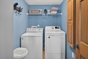 Une buanderie équipée de deux machines à laver et deux sèche-linge à chargement frontal, d’un petit panier à linge et d’une étagère sur laquelle se trouvent un autre panier et un fer à repasser.
