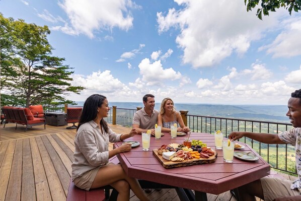 Un groupe de personnes est assis à une table, d’où l’on peut admirer l’océan, une terrasse en bois et du mobilier d’extérieur.