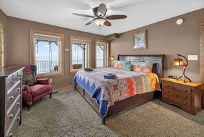 A bedroom with a bed, dresser, chair, nightstand, lamp, ceiling fan, windows, and artwork.