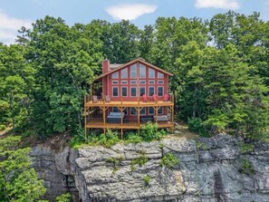A cabin with a wooden deck and a large windowed front section, surrounded by dense forest.