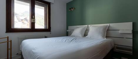 Un dormitorio con cama, ventana, radiador y estante con objetos.