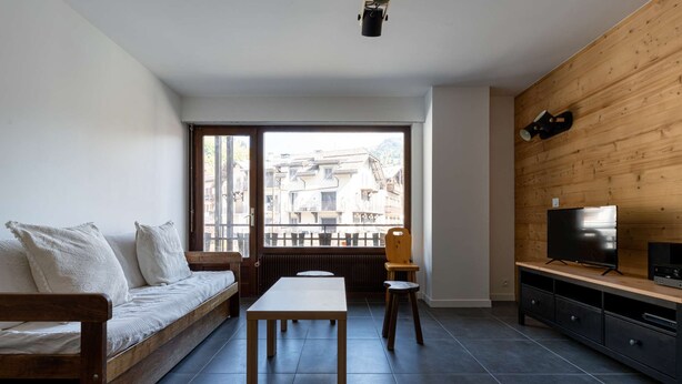 Una sala de estar moderna con un sofá, una mesa, un televisor y vistas a edificios a través de amplios ventanales.