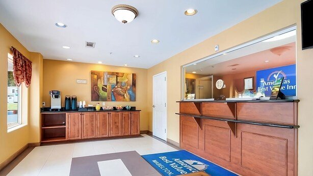 Un espace d’accueil d’hôtel avec un comptoir en bois, un coin café et un tapis bleu orné du logo de l’hôtel.