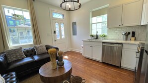 A modern living room with a black leather sofa, a wooden coffee table, and a dining area with white cabinets and a stainless steel dishwasher.