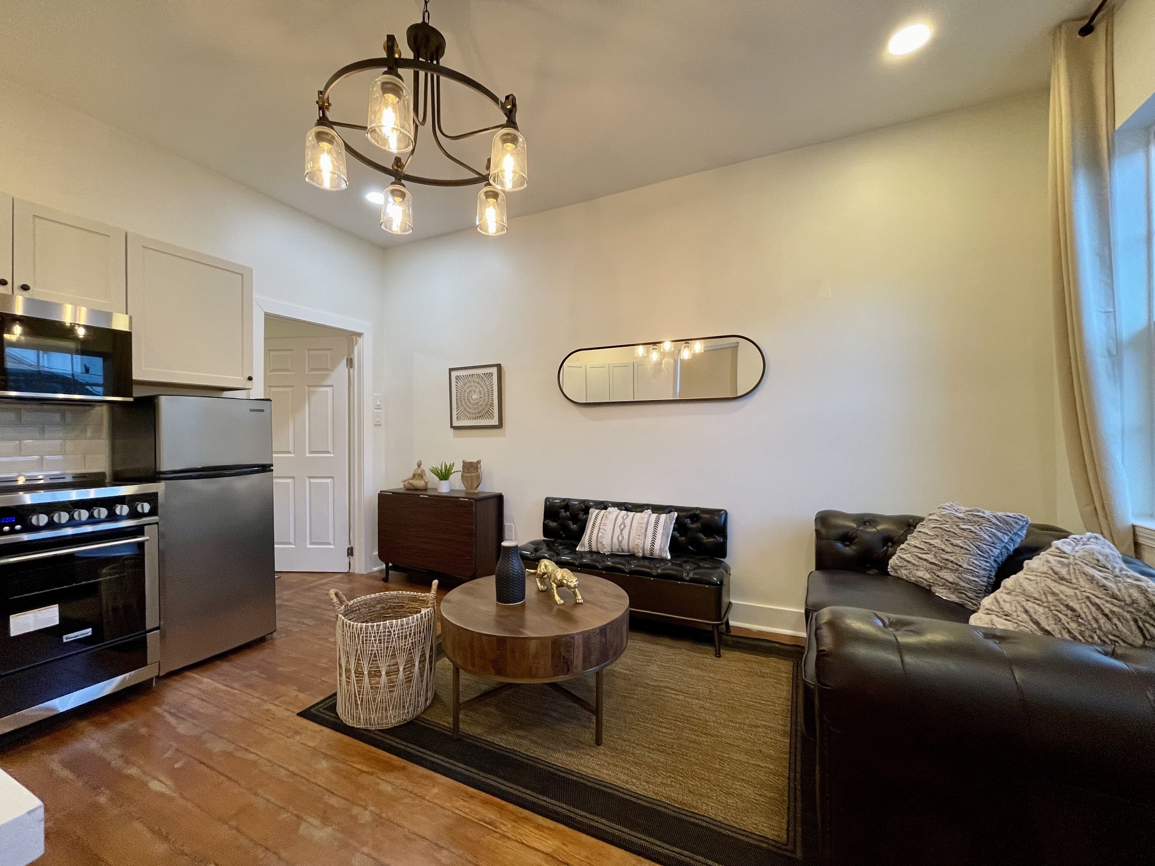 A modern kitchen with stainless steel appliances, a dining area with a round table, and a living room with a sofa and a mirror on the wall.