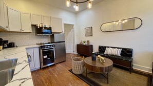 A modern kitchen with white cabinets, stainless steel appliances, and a marble countertop. There is a black leather sofa and a wooden coffee table in the living area.